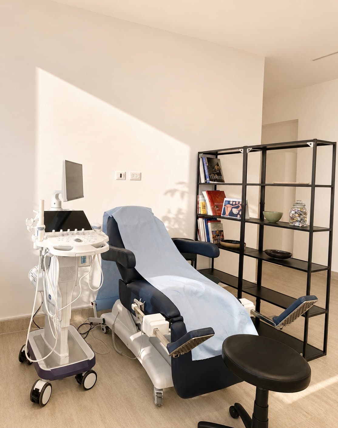 Échographe et son écran à côté d'un fauteuil d'examen recouvert d'un drap bleu, étagère de livres contre le mur.